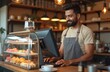© Vadym - Indian barista or owner using computer cashbox in store checking client order. Waiter in apron works in coffee shop. Small business, people and service industry. Barkeeper accepts payments, smiles.