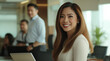 © Haram - Confident businesswoman in professional attire, smiling at her desk with a laptop while team members collaborate in a modern office with bright lighting, large windows, and a productive atmosphere.