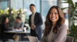 © Haram - Confident businesswoman in professional attire, smiling at her desk with a laptop while team members collaborate in a modern office with bright lighting, large windows, and a productive atmosphere.
