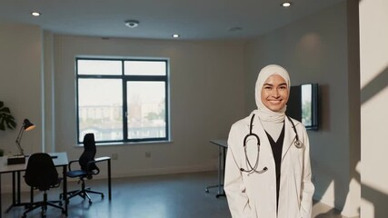 Poster - Young female muslim doctor in hijab and white lab coat confidently smiling, standing in modern medical office with bright natural lighting and professional demeanor