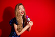 © deagreez - Smiling woman in blue sequin dress holding a pink microphone against vibrant red background, expressive and full of joy