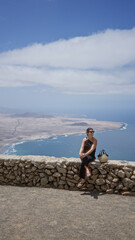 Wall Mural - Woman sightseeing outdoors in lanzarote canary islands sitting on stone wall with ocean and landscape view in bright sunny weather