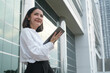 © Bordinthorn - Confident businesswoman in a white shirt holding a tablet, standing outside a modern office building, looking optimistic and forward-thinking.