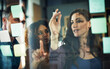 © JamDay/peopleimages.com - Business people, meeting or pointing with glass board for project planning, brainstorming or schedule at office. Colleagues, team or delegation planner with group for ideas, collaboration or vision