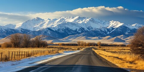 Aufkleber - Scenic mountain landscape featuring snow-capped peaks under a blue sky beside a winding road framed by golden grass and bare trees.