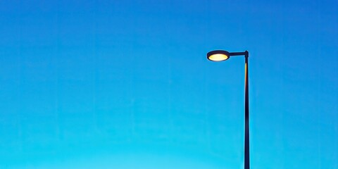 Poster - Street lamp silhouetted against a clear blue sky at twilight with warm light illuminating space below, featuring dark tones contrasting with vibrant background.
