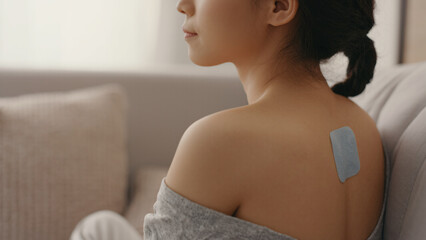 A woman with a pain relief patch on her upper back sits on a couch, wearing an off-shoulder sweater in a softly lit room.