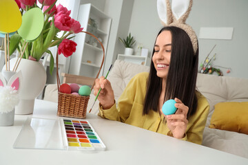 Wall Mural - Young woman in bunny ears painting Easter egg at home