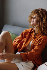 Wall Mural - Young woman with curly hair wearing an orange shirt and white shorts, sitting on a couch with a relaxed smile, embodying joy and comfort at home