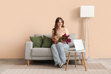 Wall Mural - Pretty young woman with bouquet of beautiful purple tulips sitting on sofa near beige wall