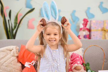 Wall Mural - Cute little girl in bunny ears with chocolate Easter eggs sitting on sofa at home