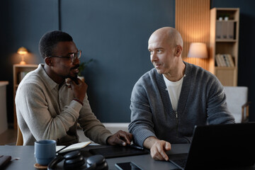 Poster - Smiling mature bald man in professional discussion with Black male colleague working collaboratively on interior design at desk with laptop and graphic tablet in office