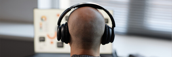Poster - Website header shot of bald man shot from rear view listening to podcast through headphones while using computer in office, copy space