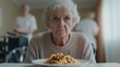 © Blackmint Studio - An elderly woman with white hair looks pensively at a plate of pasta in a care facility. Other staff members are seen in the background tending to residents during the afternoon