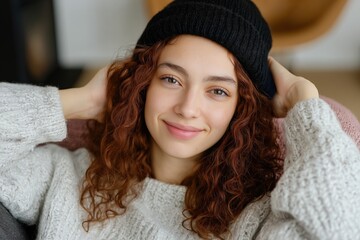 Wall Mural - A woman with red hair is wearing a black hat and smiling. She is sitting on a couch and she is relaxed