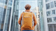 © Number One Studio - person with curly hair wearing orange sweater and yellow backpack stands in front of modern glass building, reflecting urban life and adventure