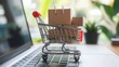 © MichaelMob - A close-up of a shopping cart filled with small cardboard boxes placed on a laptop keyboard, symbolizing online shopping and e-commerce in a modern setting with greenery.