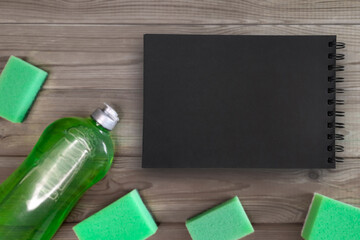 Wall Mural - Blank open notebook with black pages on grey wooden blurred background with green cleaning items. Template with place for text, Top view, Copy space for Planning housekeeping cleaning.