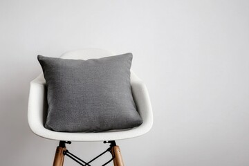 Poster - A gray pillow rests on a white chair against a white wall