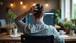 © TheWaterMeloonProjec - Contemplating innovative ideas. Back view of a young man in casual attire with his hand resting on the back of his head, working at a desk in a creative workspace.