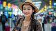 © Imagine  - A cheerful young woman with a straw hat and braided hair, using her smartphone and wearing earphones in a busy airport terminal, enjoying travel and staying connected on her journey