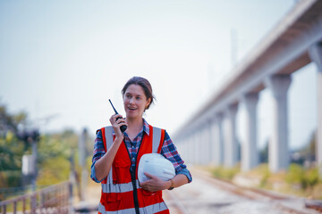 Wall Mural - White female Rail transportation engineer in safety vest and hardhat using walkie-talkie at the railway track with moterway background.