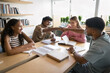 © fizkes - Diverse group of motivated students engaged in productive learning in library seated around desk with open books and notebooks, focus on two guys giving each other fist bump, symbol of teamwork, unity