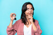 © luismolinero - Young hispanic woman holding home keys isolated on blue background having doubts while looking up