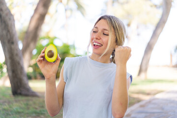 Wall Mural - Young blonde woman holding an avocado at outdoors celebrating a victory