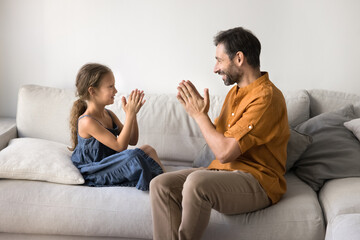 Middle-aged loving father and beautiful preschooler daughter spend playtime on leisure together at home, sitting on comfortable couch, playing patty-cake. Family games, interaction and joyful pastime