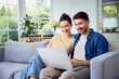 © baranq - Young couple sitting on sofa at home, smiling while using laptop in living room