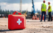© AbsoluteAi - Construction site worker safety and first aid kit. A red first aid kit is placed on a construction site, with workers in the background wearing safety vests and helmets.