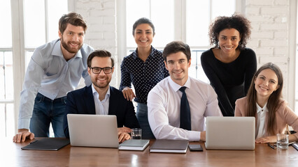 Canvas Print - Six business professionals posed together at table with laptops in meeting room or workspace. Team members smiling at camera, looking confident and engaged in positive, productive, united team work