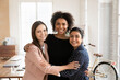 © fizkes - Three beautiful multinational women colleagues, team mates posing indoors, hugging, standing in modern creative workspace looking at camera. Racial equality, friendship at work, teamwork and synergy