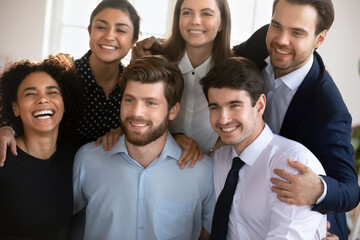 Canvas Print - Group of young attractive workmates, multiethnic team members hugging indoors, smile look aside, showing camaraderie, strong sense of unity and togetherness. Friendship at work, synergy and support