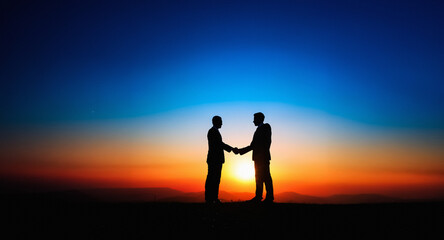 Aufkleber - Two silhouetted figures shake hands against a striking sunset backdrop. The sky blends from deep blue to vibrant orange and red as the sun dips below the horizon, creating a symbolic scene of agreemen