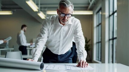 Poster - Focused mature male engineer in white shirt and glasses analyzing blueprints on a white table in a modern office, captured in a slow motion sequence
