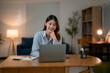 © Parichat - Cheerful Asian professional typing on laptop, sitting at wooden desk in dimly lit living room, wearing casual attire and smiling while focusing on work screen