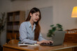 © Parichat - Focused female professional working late, typing on laptop at home office, surrounded by notes, reference books, soft lamp illumination highlighting workspace