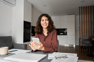 Canvas Print - Smiling dreaming young latin hispanic woman using online smartphone app remote communication, doing payment for shopping or reading news at home. Mid age businesswoman working at mobile phone device