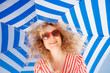 © Sunny studio - Happy young woman under a striped blue parasol. Girl wearing sunglasses enjoys a summer vacation