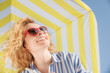 © Sunny studio - Happy young woman under a striped yellow parasol. Girl wearing sunglasses enjoys a summer vacation