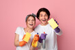 © Prostock-studio - Happy couple holding spray detergents and cleaning sponges and looking at camera, posing over pink background in studio, free space