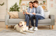 © Prostock-studio - Leisure Time. Portrait of young loving family sitting on couch, using digital tablet, happy labrador lying on the floor. Married woman and man spending time together at home in living room, holding pc