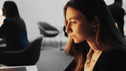 Aufkleber - Office worker concentrating on her laptop, occasionally looking away thoughtfully, while colleagues discuss in the background of a brightly lit modern office