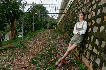 Wall Mural - Young woman in casual outfit enjoying nature in the garden, showcasing a serene expression and a stylish look with a green backdrop