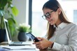 © velikiyzayats - Young Adult Woman in Business Attire Using Smartphone While Sitting at Desk with Laptop and Green Plant in Modern Office Environment