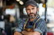 © SY - A mature man with a beard, wearing a plaid shirt and apron, stands in a workshop with arms crossed. This image portrays skilled labor, craftsmanship, or manual work.
