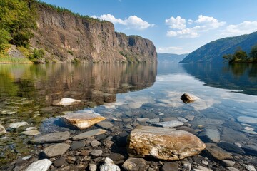 Aufkleber - Tranquil mountain lake with rugged cliff reflection and clear water with stones under blue sky and fluffy clouds