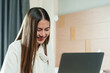 © Ekkasit A Siam - Asian woman in a white blouse smiling while working on a laptop in a cozy home. happiness and enjoyment in remote work, freelancing, or online business, reflecting a modern and flexible lifestyle.